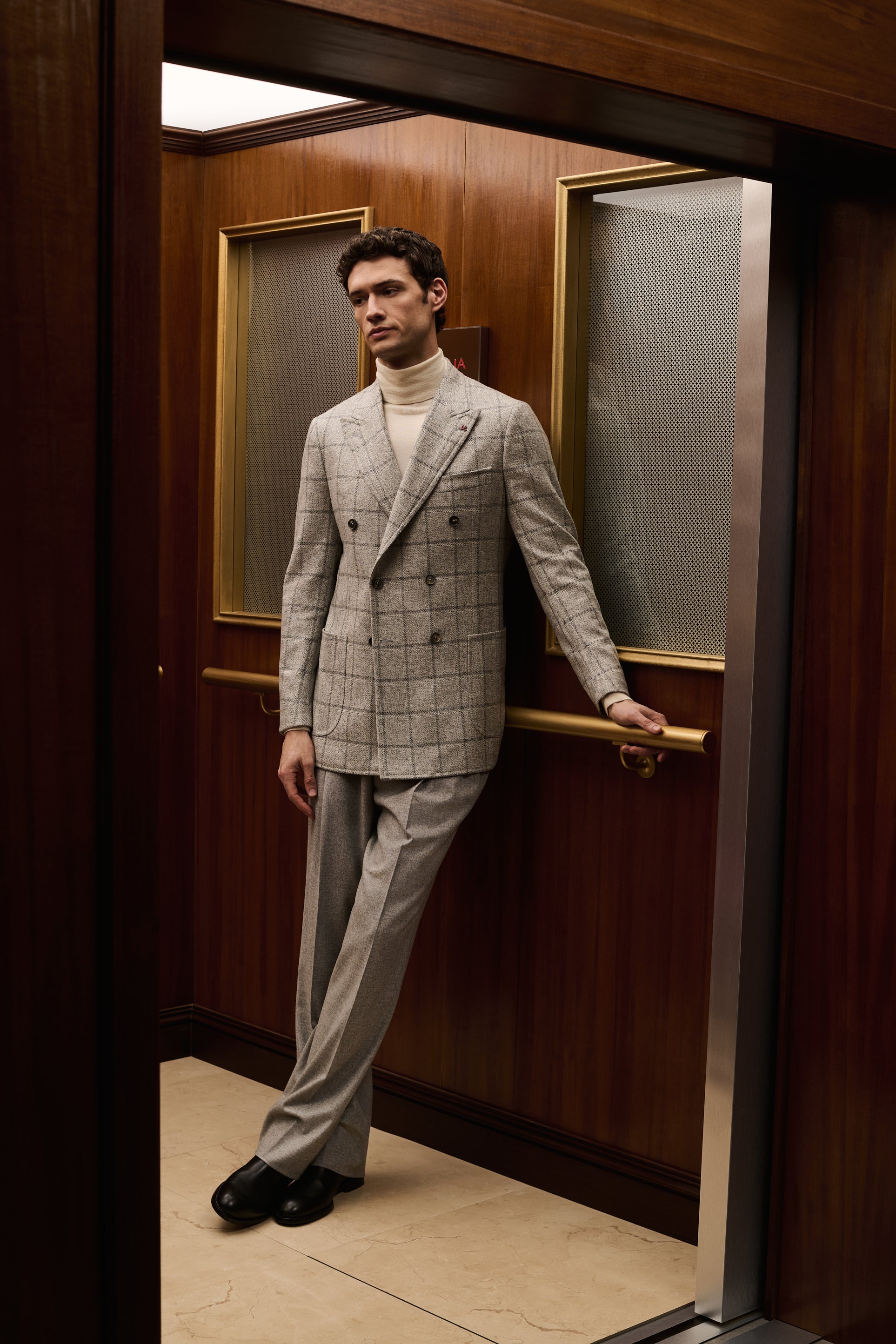 ISAIA Made to Measure: light grey check double-breasted suit with cream rollneck, leaning on the elevator handrail