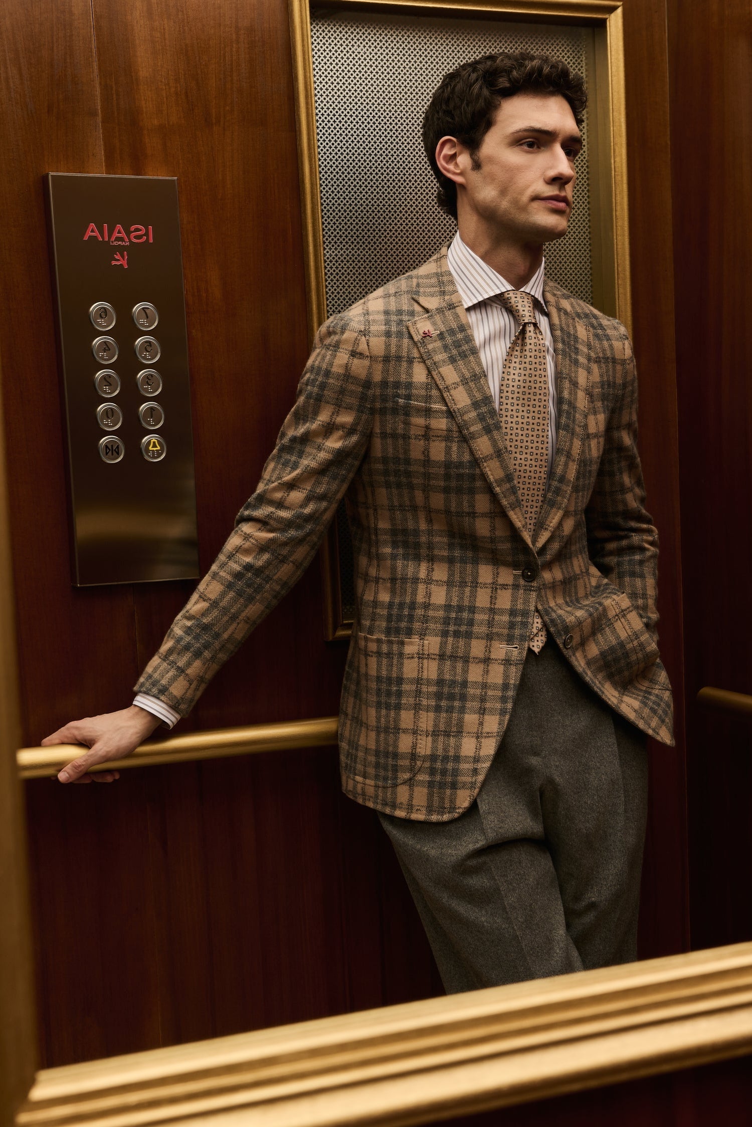 ISAIA Made to Measure: camel check jacket with beige tie and grey trousers, leaning on the elevator handrail