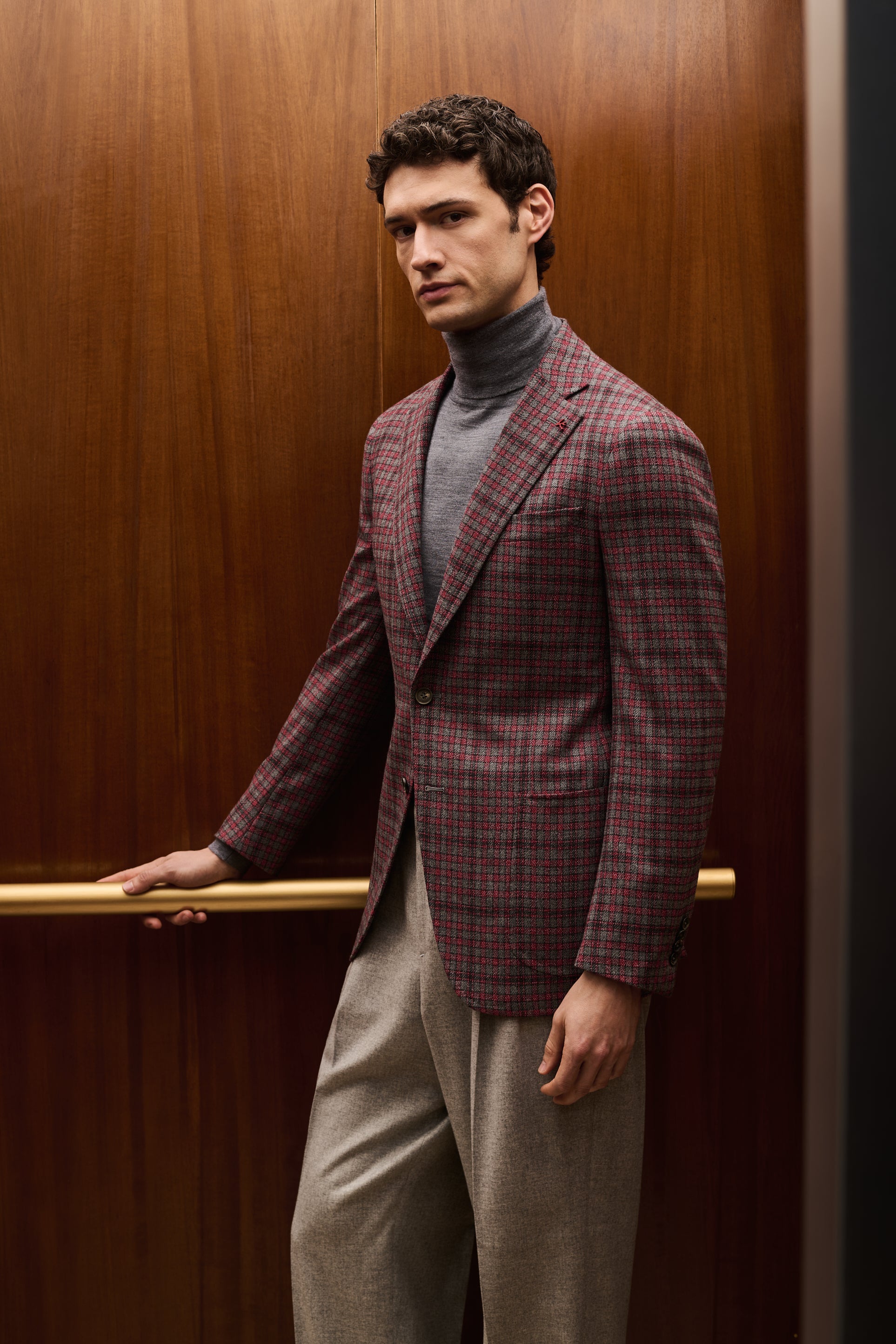 ISAIA Made to Measure: plum check jacket with grey rollneck and grey trousers, a hand on the elevator handrail