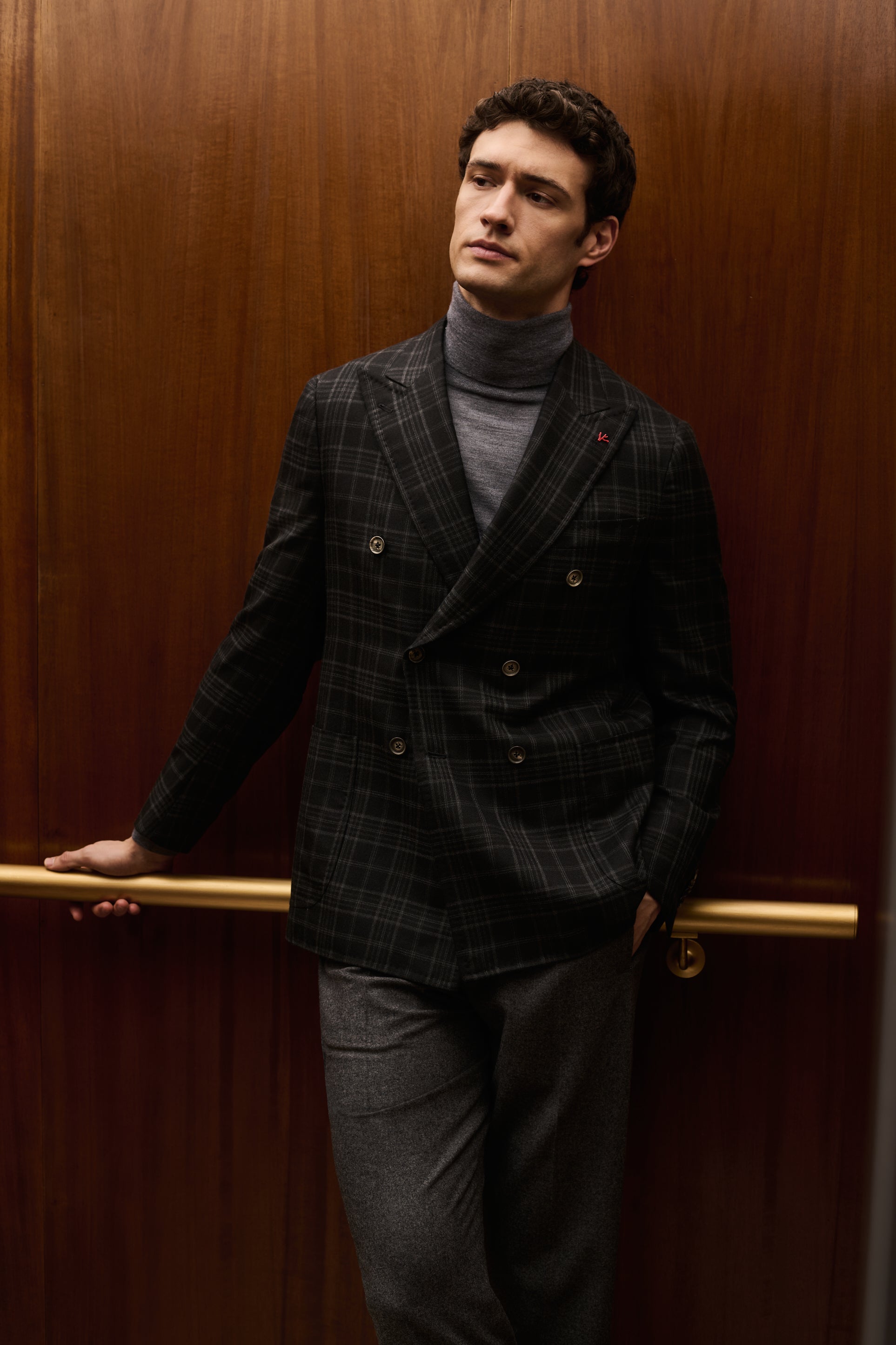 ISAIA Made to Measure: dark check double-breasted jacket with grey rollneck, leaning on the elevator handrail