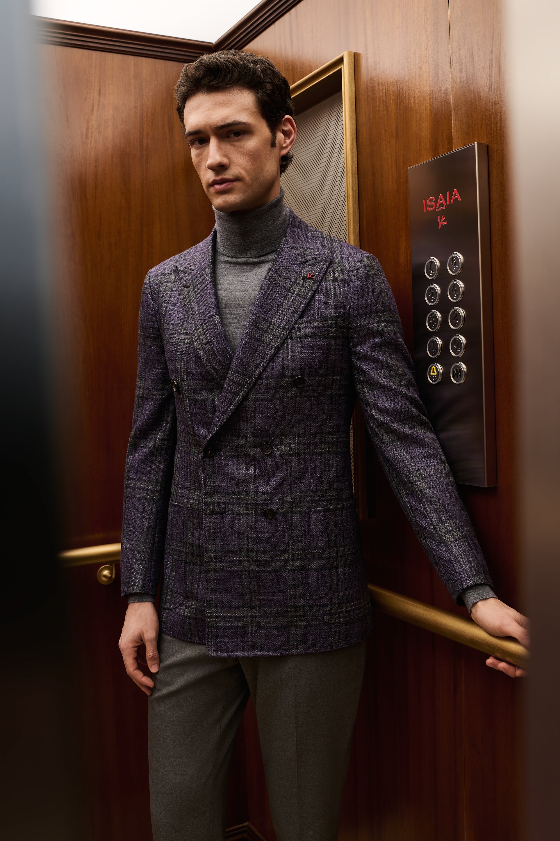 ISAIA Made to Measure: purple check double-breasted jacket with grey rollneck, holding the elevator handrail