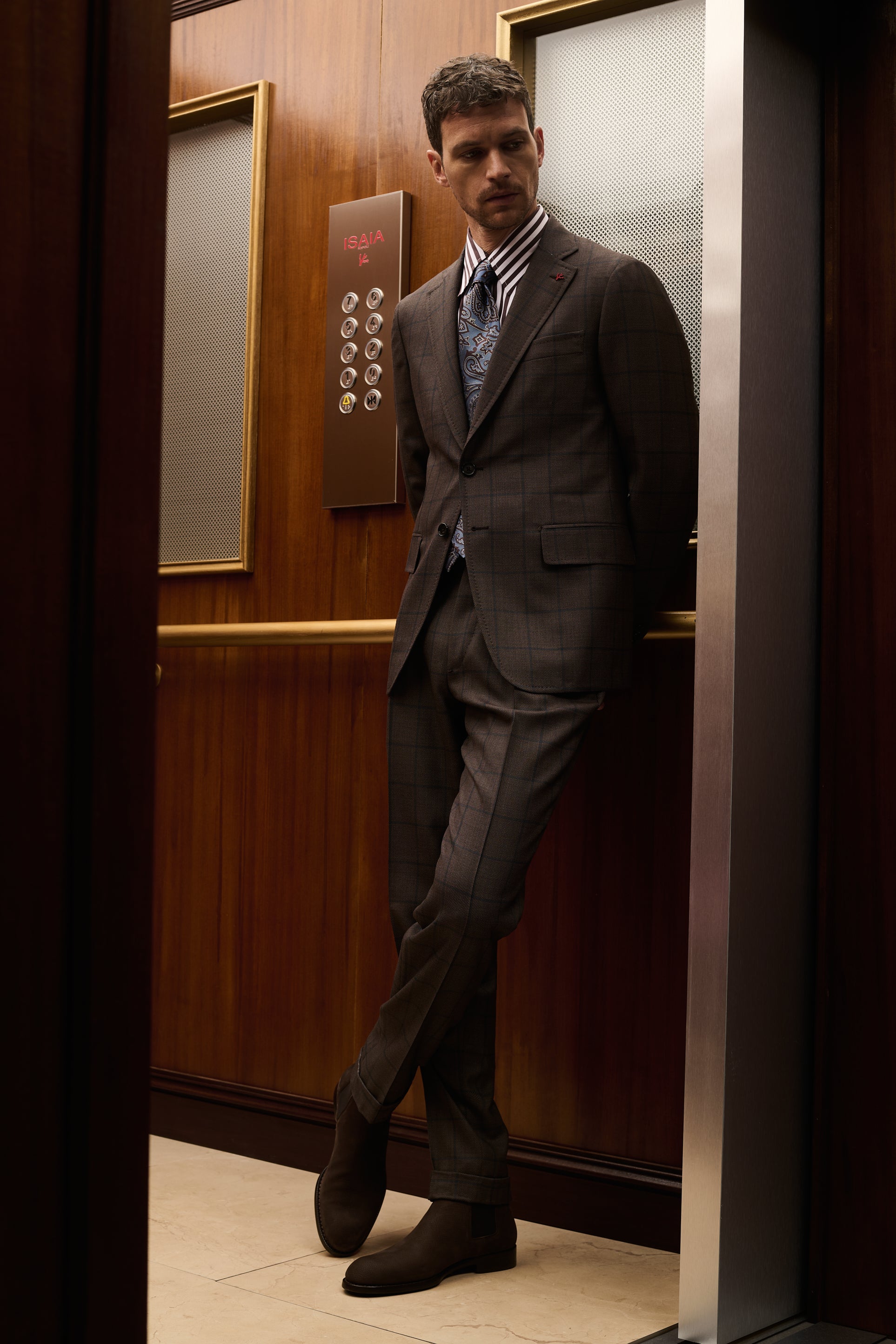 ISAIA Made to Measure: brown suit with striped shirt and patterned tie, leaning against the elevator door