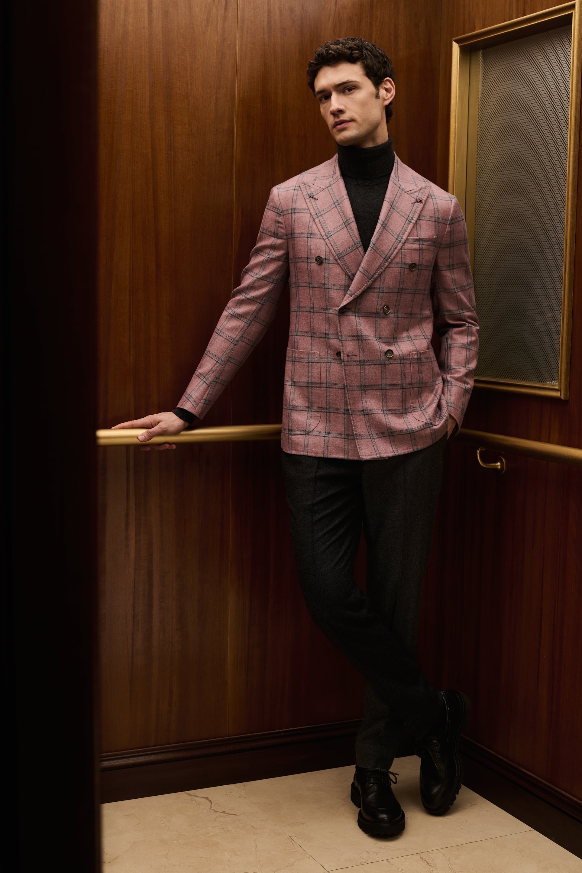 ISAIA Made to Measure: pink check double-breasted jacket with black rollneck, leaning on the elevator handrail