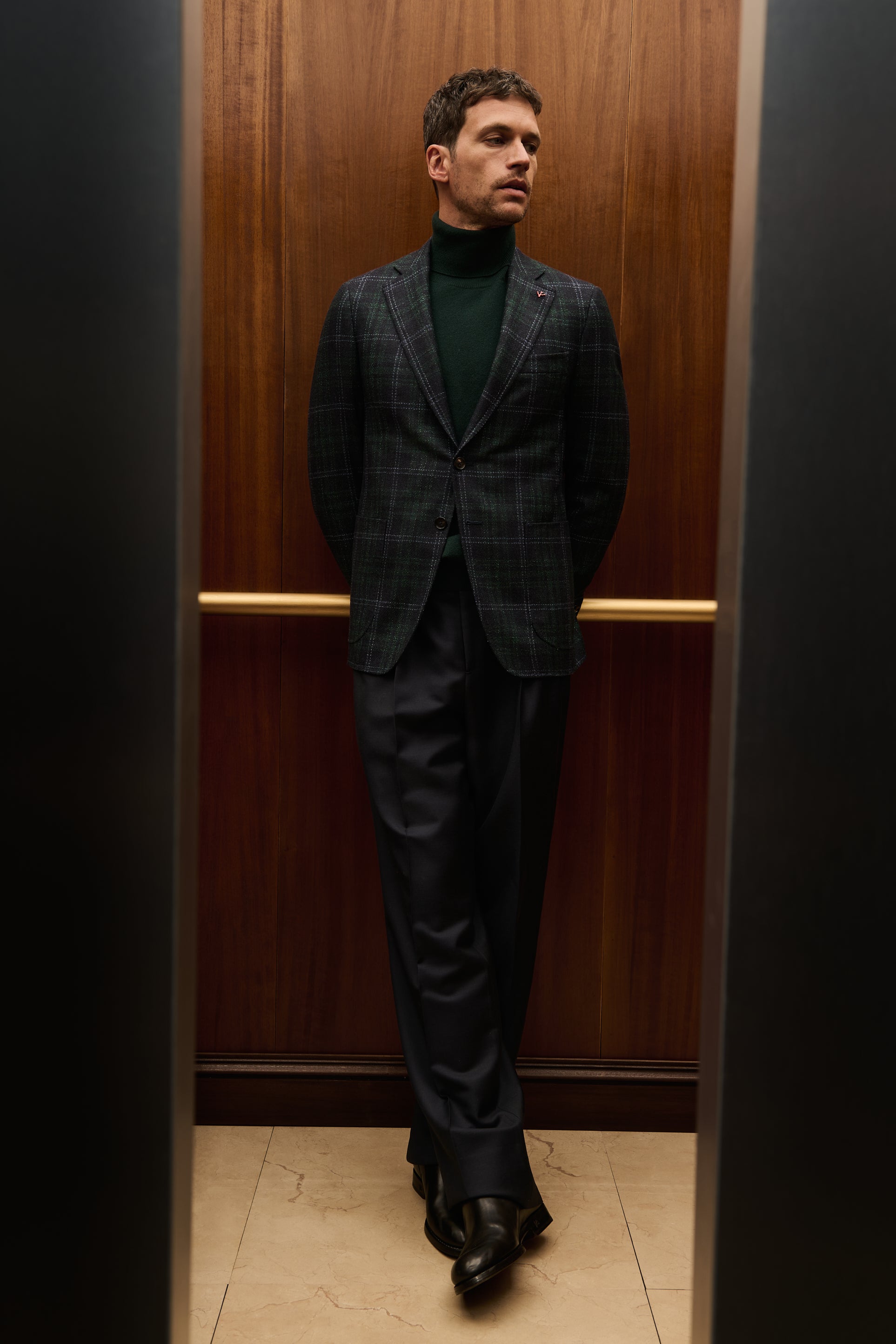 ISAIA Made to Measure: grey check suit with dark green rollneck, leaning against the elevator wall